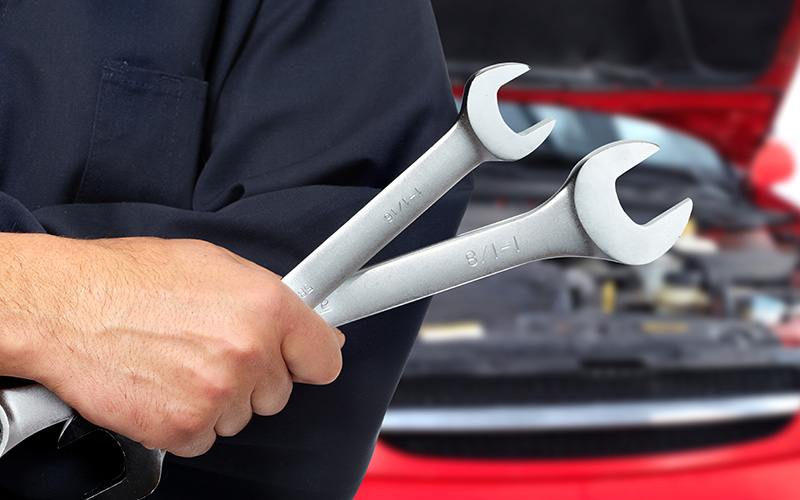 Automotive technician holding two wrenches in front of an open car hood, highlighting professional vehicle maintenance and repair services.