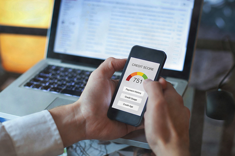 A close-up of a person holding a smartphone that displays a credit score of 751 with a colorful dial gauge, with a blurred laptop in the background.