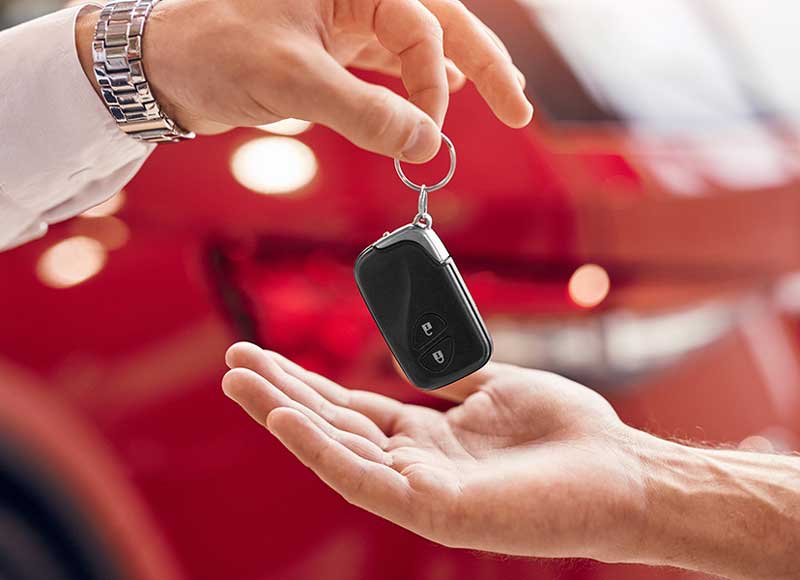 A close-up of one person's hand dropping a black car key fob into another open hand, with a blurred red car in the background.